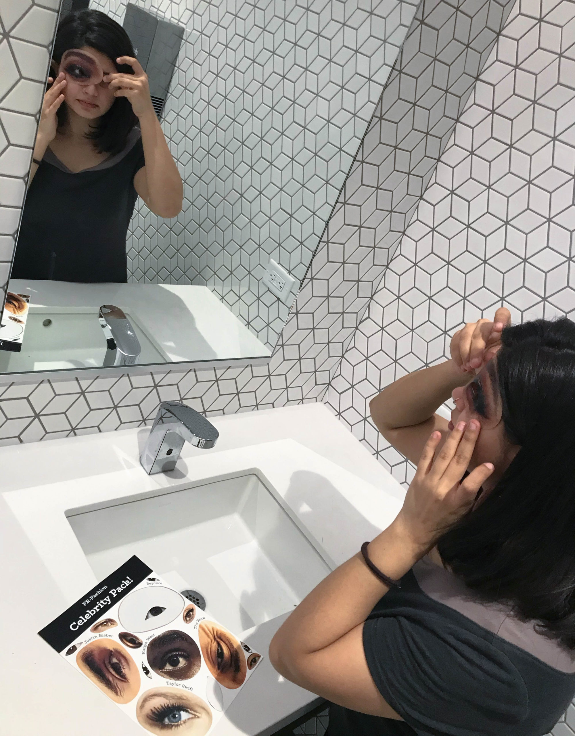 and Indian woman looking in the mirror of a bathroom mirror as she puts a sticker with a cut-out over her eye. A sheet of stickers is next to her on the counter