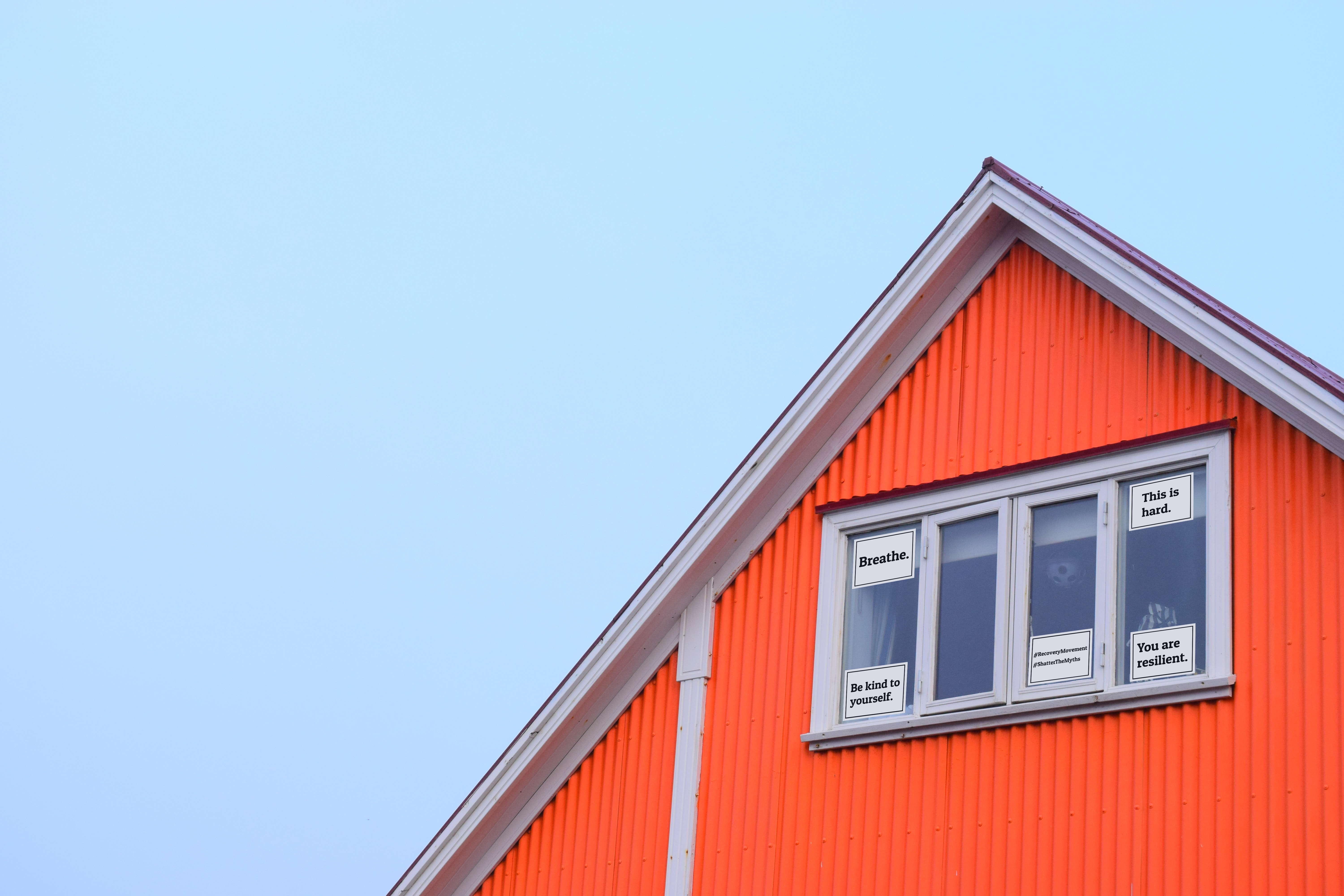 a red house with a white framed window. Five signs in the window read: "Breathe.", "This is hard.", "Be kind to yourself." "You are resilient.", and "#RecoveryMovement"