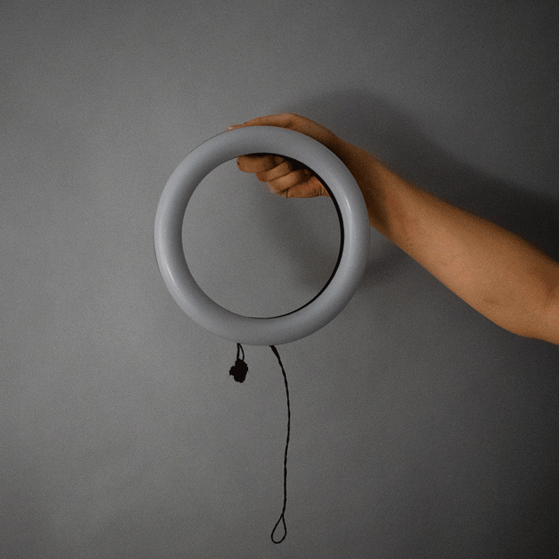 gif of a ring light being held up by a human arm. Three frames in the gif show a soft carbon fiber switch hanging loose as a string and a ball, with the switch about to be connected, and with the switch connected and the light shining