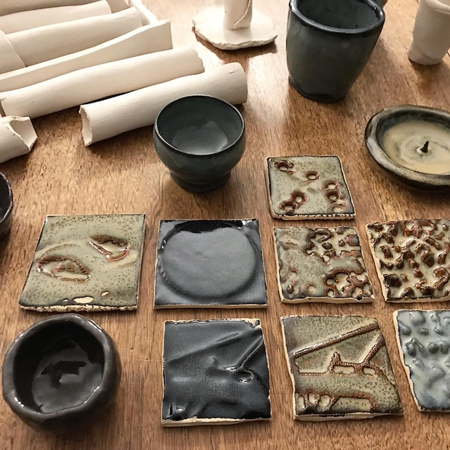 Ceramic samples on a wood table: 8 glazed tiles, 4 glazed vessels, various unglazed vessels in the background