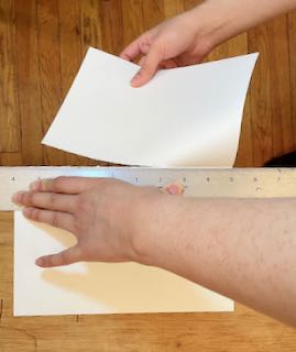 light skinned hands ripping a sheet of watercolor paper in half on the edge of a table with a metal ruler holding the edge down to the table