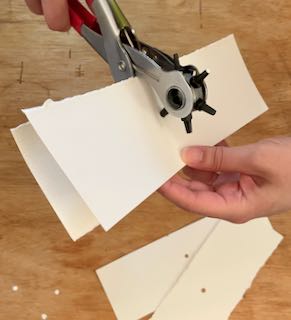 light skinned hands punching a hole in the center of rectangular pieces of watercolor paper with a leather punch. A previously punch sheet is lined up with an unpunched sheet to make the new hole align with the old
