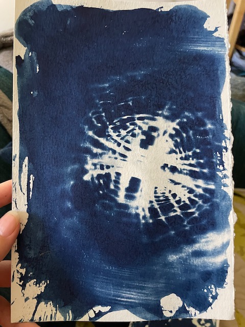 a hand holding up a cyanotype contact of a woven basket, the basket bottom is visible and the sides gently fade away so that the print looks like a watery spiderweb