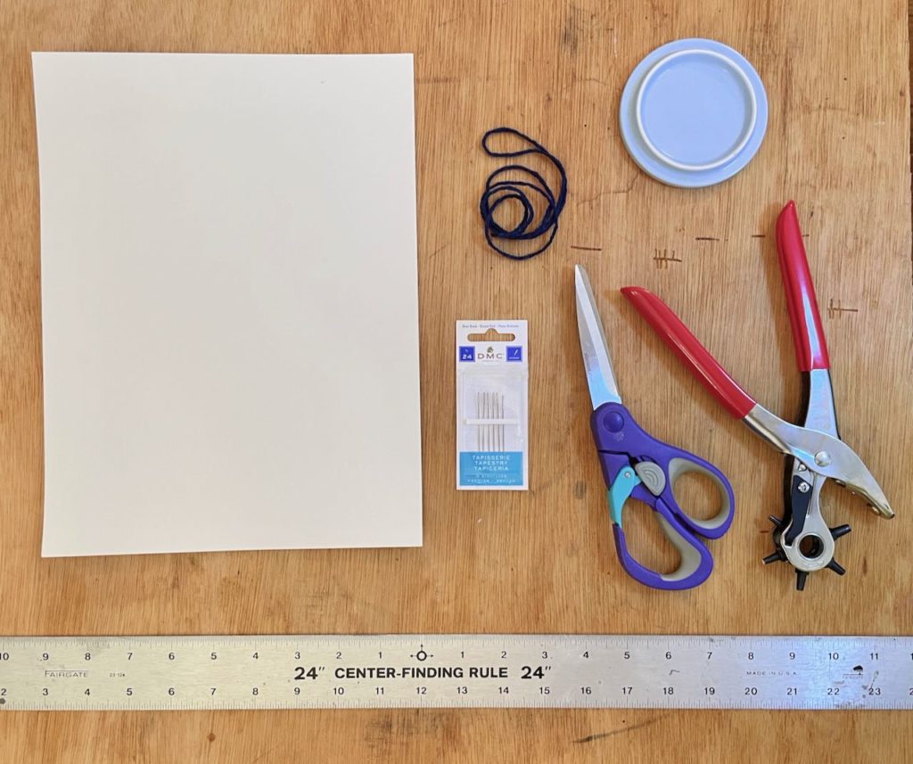 From top left to bottom right: white watercolor paper, dark blue yarn, ceramic mug lid, metal tapestry needles, scissors, leather punch, metal ruler