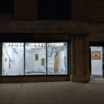 a view of the exhibition from outside. it is dark out but the lights are on, making the brown chromatograms and blue streamers glow